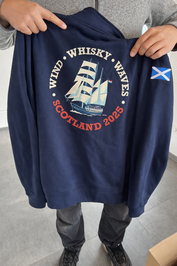 Rückseite eines Sweatshirt mit runden Emblem "Wind - Whisky - Waves". In der Mitte ist eine Illustration eines Segelschiffs.