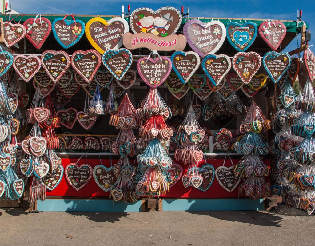Foto eines Lebkuchenherzerlstands auf dem Münchner Oktoberfest. Eines der großen Lebkuchenherzen oben am Stand ist aus Holz, mit zwei Comic-Herzen die sich verliebt anblicken. Ergänzt wird das Holzherz durch ein geschwungenes Schild mit den Schriftzug " A nettes Herzl".